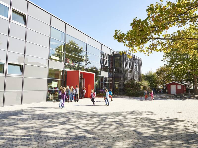 Ein moderner Schulhof mit spielenden Kindern vor einem großen, grauen Gebäude mit roten Elementen und Bäumen im Hintergrund.