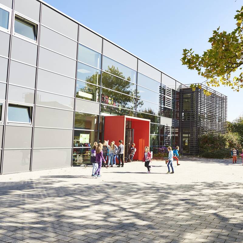 Ein moderner Schulhof mit spielenden Kindern vor einem großen, grauen Gebäude mit roten Elementen und Bäumen im Hintergrund.