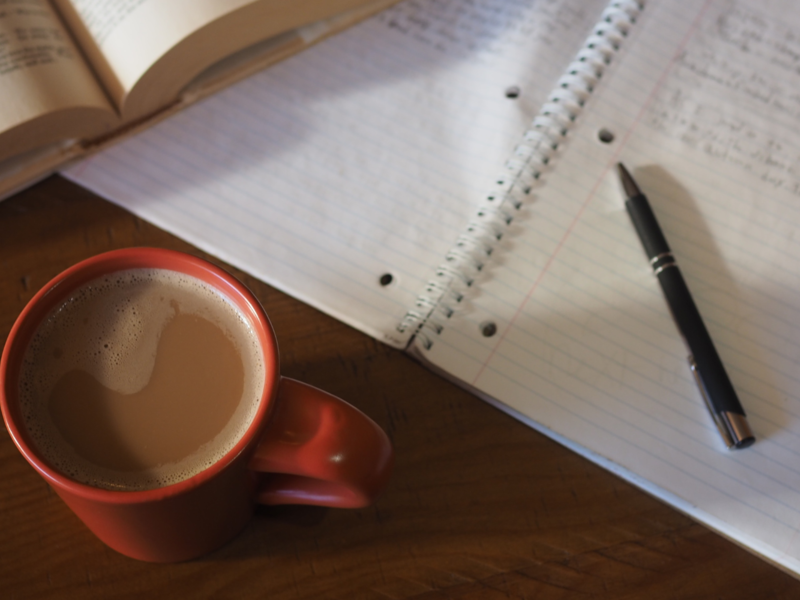 Auf einem Tisch liegt ein aufgeschlagenes Buch, ein Notizblock mit Notizen und einem Stift. Eine rote Kaffeetasse steht davor.