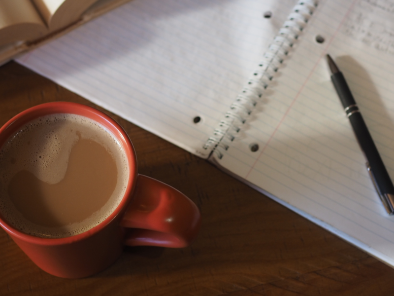 Auf einem Tisch liegt ein aufgeschlagenes Buch, ein Notizblock mit Notizen und einem Stift. Eine rote Kaffeetasse steht davor.