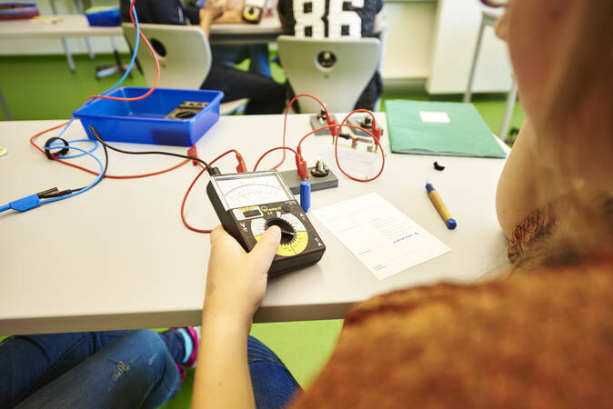 Ein Arbeitstisch mit aufgebauten technischen Versuchen darauf. Man blickt einer Person, die einen Test durchführ, über die Schulter.